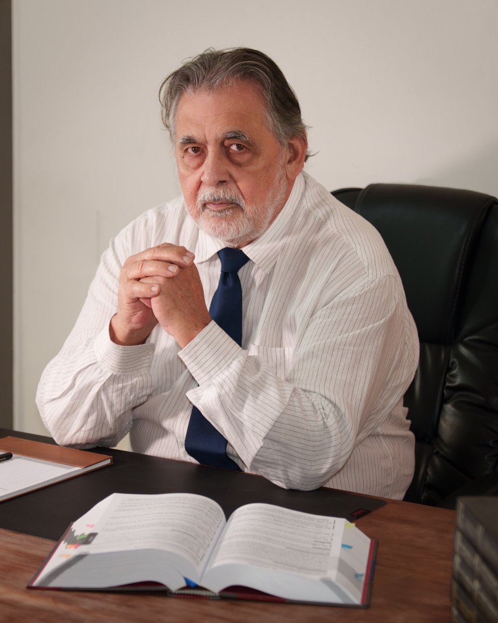 Arlindo Victor sentado à mesa do escritório, com as mãos entrelaçadas e um livro de direito aberto à frente